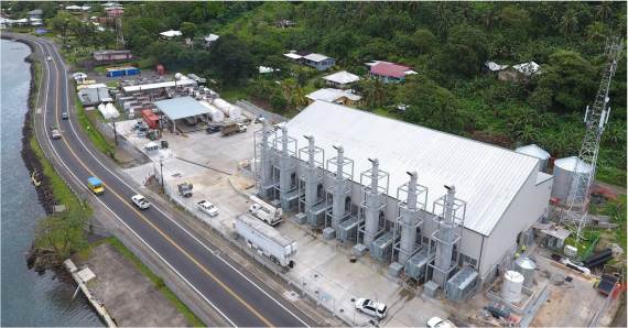 Aerial view of a coastal industrial facility in American Samoa near a road with multiple vehicles parked. The facility, crucial for storing lubricant oil and fuel, features a large building with several ventilation shafts and surrounding equipment. The road runs parallel to the shoreline on the left, with lush green vegetation around.