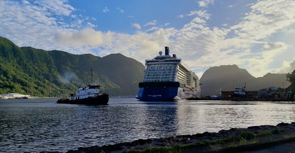 A large cruise ship docks at a port in American Samoa, surrounded by lush, green mountains and a partially cloudy blue sky. A smaller tugboat moves near the cruise ship, assisting with its docking. The calm water reflects parts of the sky and the idyllic scenery.