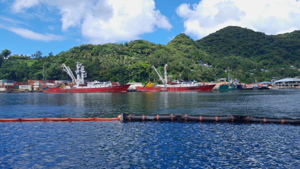 Two red fishing boats are docked by a lush, green hillside in American Samoa. A village with houses is partially visible among the trees. The blue sky is filled with scattered white clouds. In the foreground, an orange buoy and a black floating barrier on the calm water mark where fuel and lubricant oil supplies are managed.