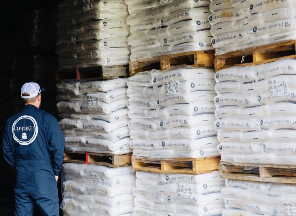 A person wearing a blue jumpsuit and white cap stands facing away, looking at stacks of large white bags on wooden pallets in a warehouse. The bags, labeled "Clipper Coarse Salt," are neatly organized.