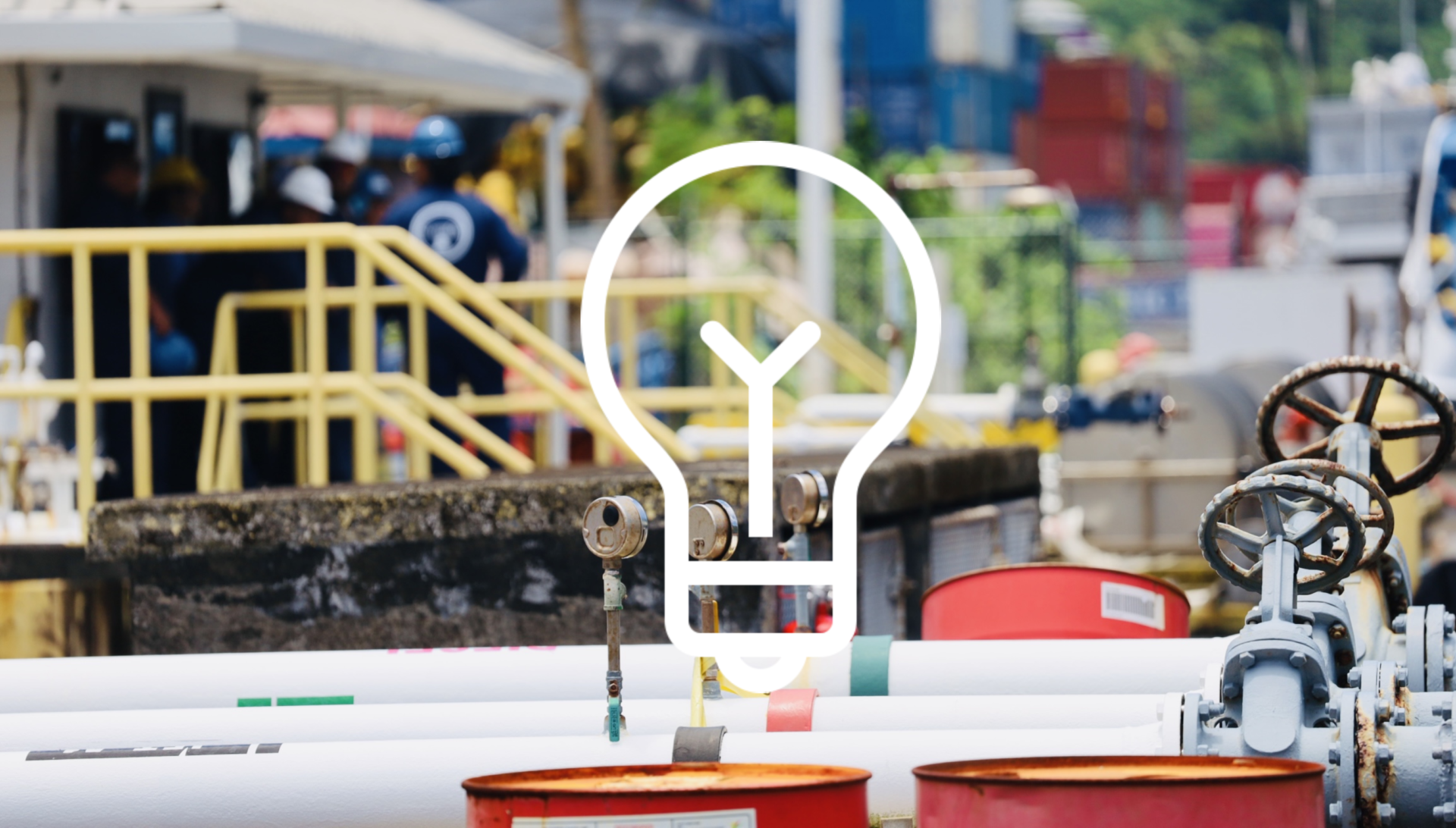 An industrial setting features workers wearing helmets and safety gear, surrounded by machinery and large pipes. Various equipment, drums, and diesel fuel containers are in the foreground. A transparent lightbulb icon is superimposed over the image, symbolizing innovation or ideas.