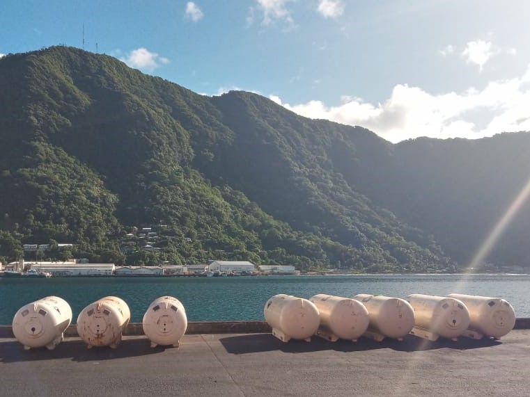 Large white cylindrical tanks lie on a dock by a blue body of water, likely storing NH3 for refrigeration solutions. In the background, a green, forested mountain range rises, partially covered in shadow under a partly cloudy sky. Small buildings are visible at the base of the mountains.