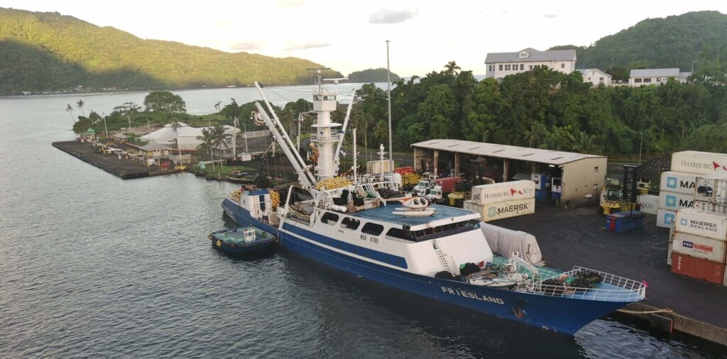 An aerial view of a cargo and research ship named "FIRE ISLAND" docked at a port. Surrounded by lush green hills and trees, the ship is being refueled as multiple containers, including those for lubricant oil, sit nearby. A few buildings dot the background under a partly cloudy sky illuminated by sunlight.